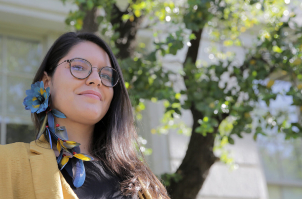 Photo of Astolfi standing in front of a soapbark tree on the UC Berkeley campus, wearing an earring of macaw feathers.